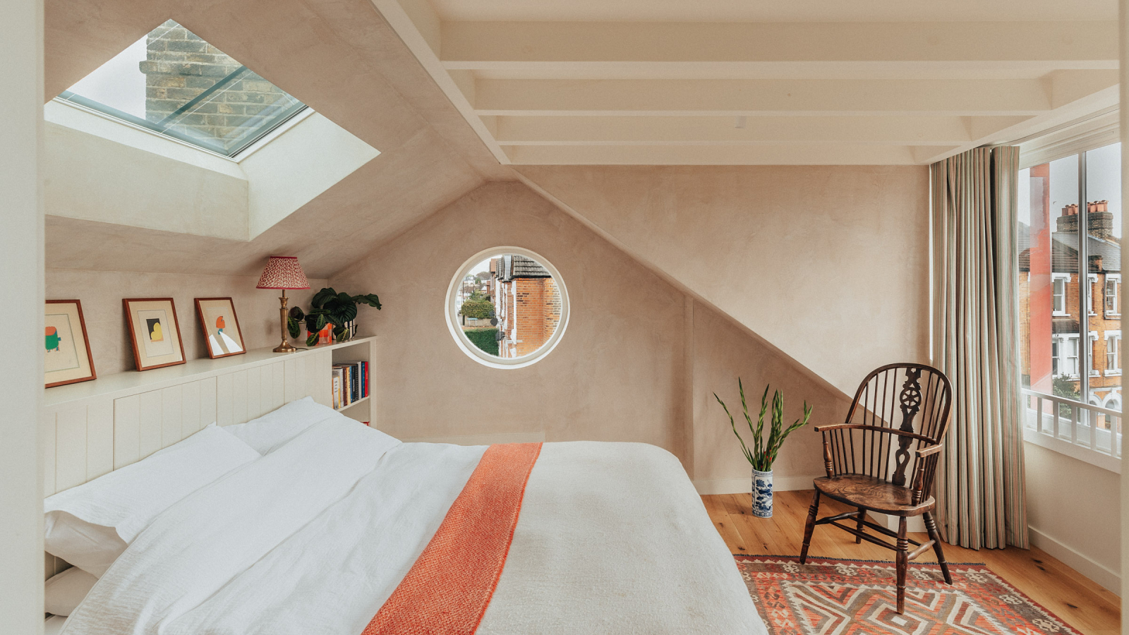 dormer loft conversion bedroom with large windows looking out, bed, built in headboard and shelving, small rooflight above bed and pale pink plaster effect walls with porthole window on one side of the room