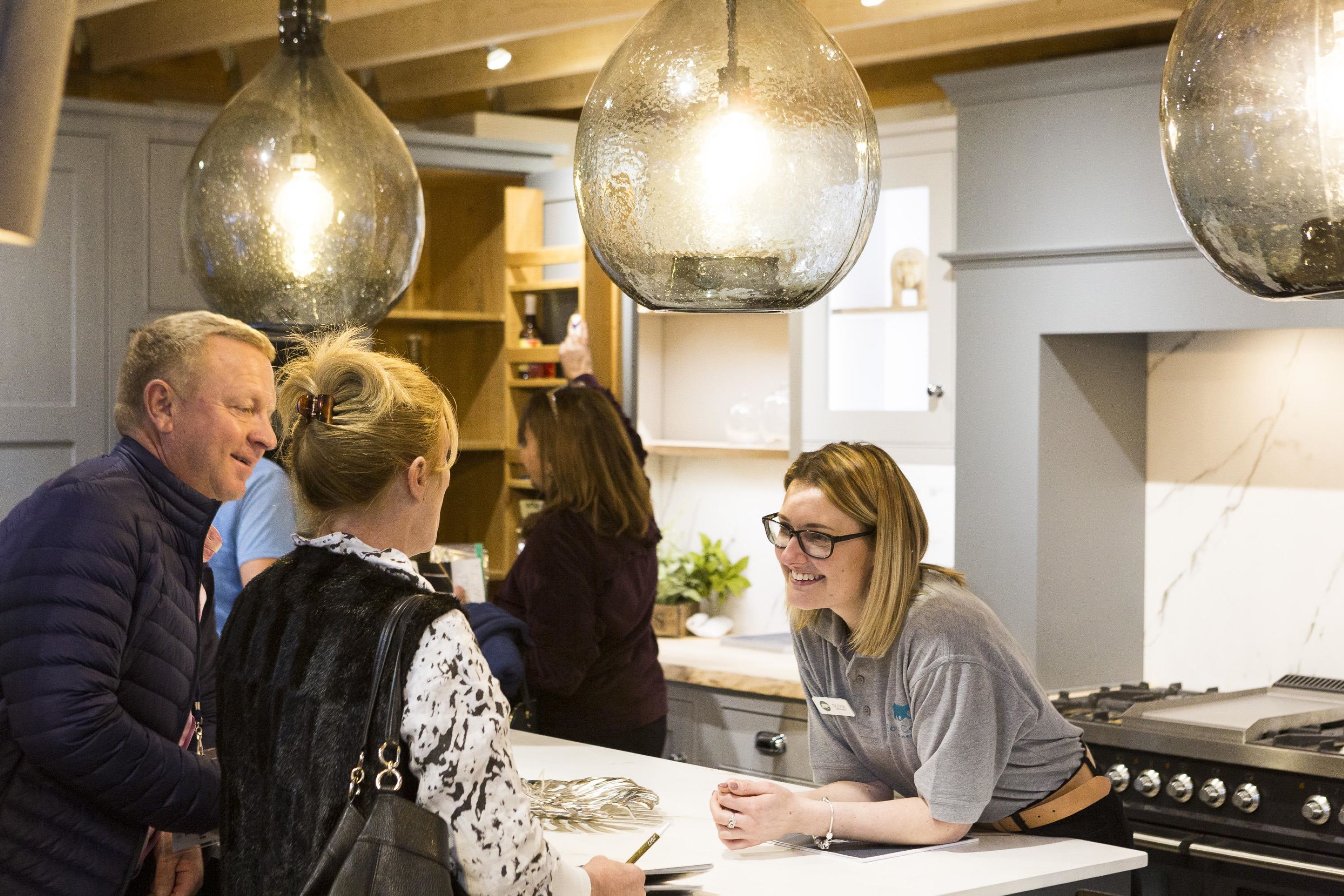 A couple at the Homebuilding &amp; Renovating Show under a bright overhead light asking a woman at a stall for some advice